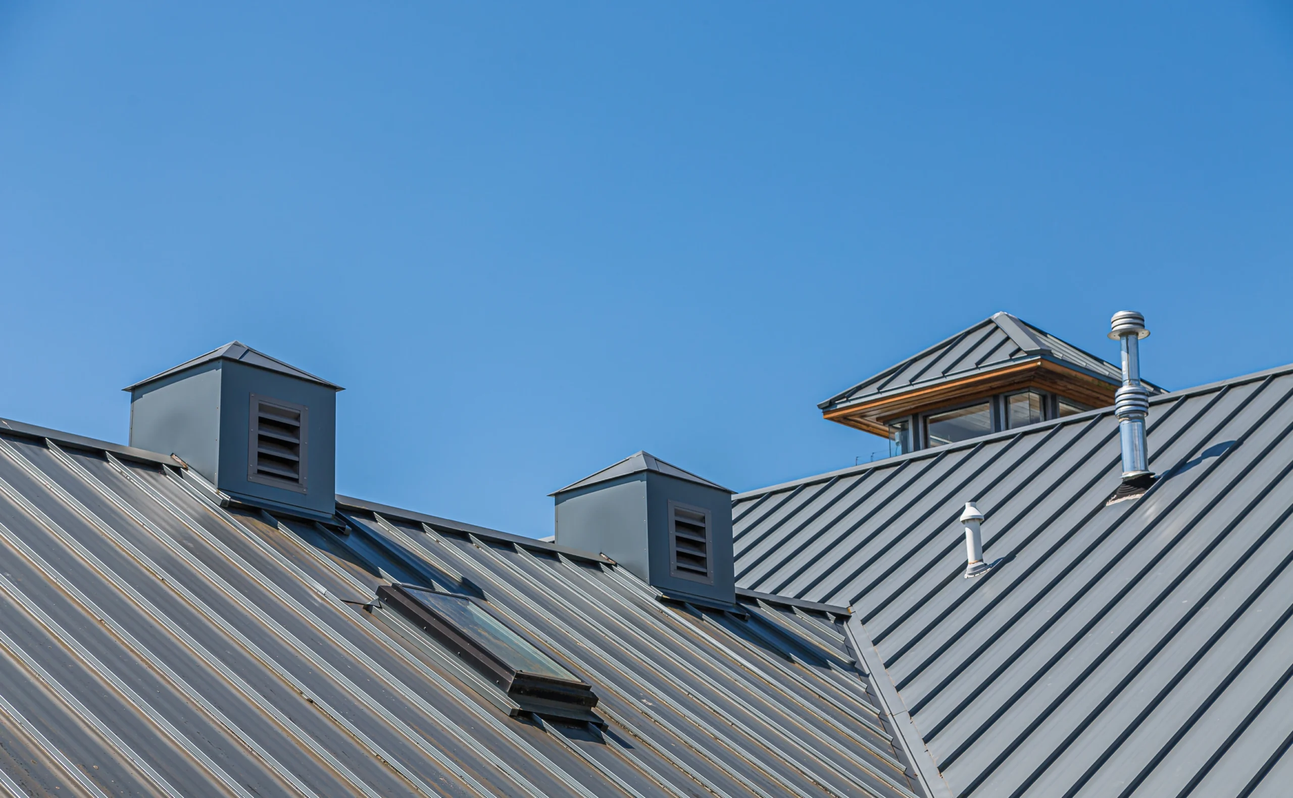 Modern metal roofing with vents, skylight, and chimney pipes under clear blue sky.